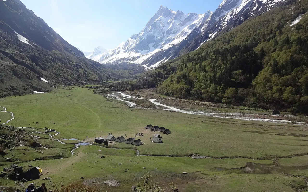 A green Himalayan valley with tents beside a river and snowy peaks beyond